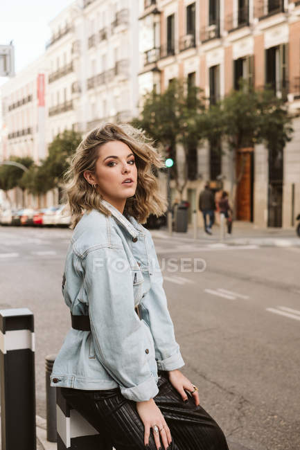 Привлекательная женщина в модном наряде сидит на болларде на городской улице — стоковое фото