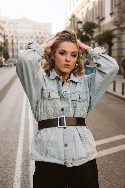 Attractive female in trendy outfit standing in middle of asphalt road touching hair on city street — Stock Photo
