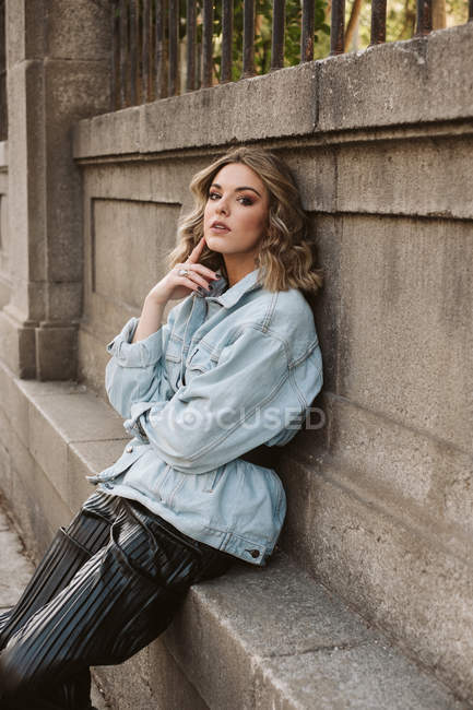 Jeune femme en tenue élégante regardant la caméra tout en étant assis près de la clôture en pierre du parc de la ville — Photo de stock