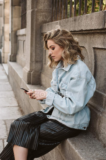 Giovane signora in elegante vestito smartphone di navigazione mentre seduto vicino alla recinzione di pietra del parco della città — Foto stock