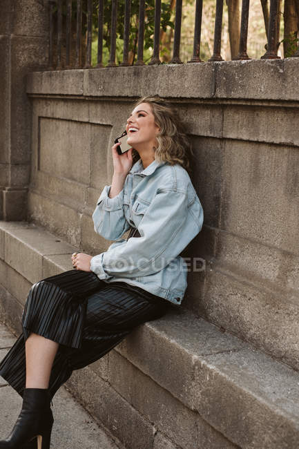 Молода леді в стильному вбранні на мобільному телефоні, сидячи біля кам'яного паркану міського парку — стокове фото