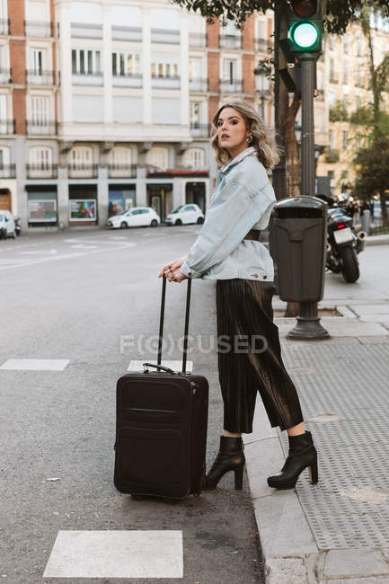 Стильна молода жінка з валізою, яка дивиться далеко під час перетину міської вулиці — стокове фото