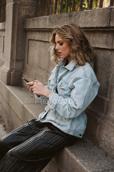 Jovem senhora em elegante equipamento de navegação smartphone enquanto sentado perto de cerca de pedra do parque da cidade — Fotografia de Stock