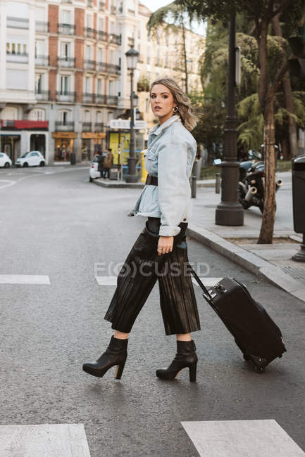 Jeune femme élégante avec valise regardant loin tout en traversant la rue de la ville — Photo de stock