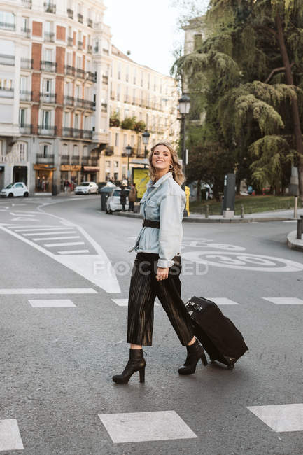 Усміхнена стильна молода жінка з валізою, яка дивиться далеко під час перетину міської вулиці — стокове фото