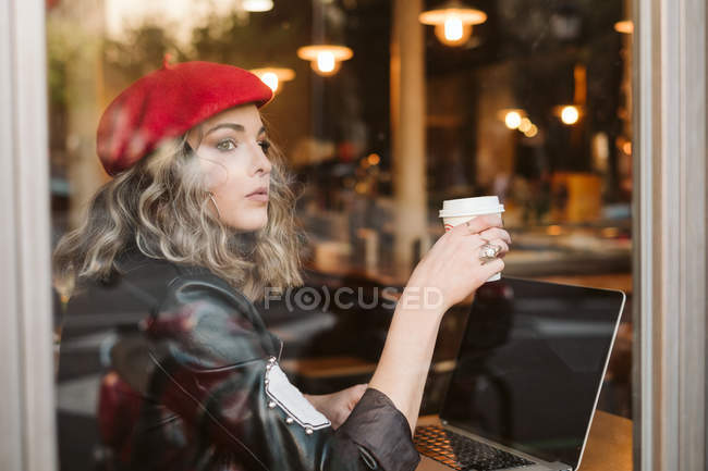 Jovem fêmea em boina vermelha bebendo bebida quente e olhando para fora da janela enquanto navega laptop no café — Fotografia de Stock