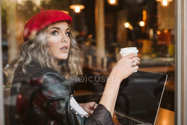 Junge Frau in roter Baskenmütze trinkt Heißgetränk und schaut aus dem Fenster, während sie im Café Laptop surft — Stockfoto