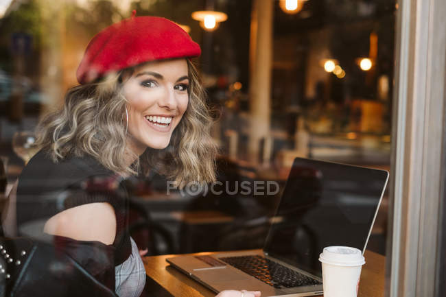 Веселая молодая женщина в красном берете пьет горячий напиток и смотрит в окно во время просмотра ноутбука в кафе — стоковое фото