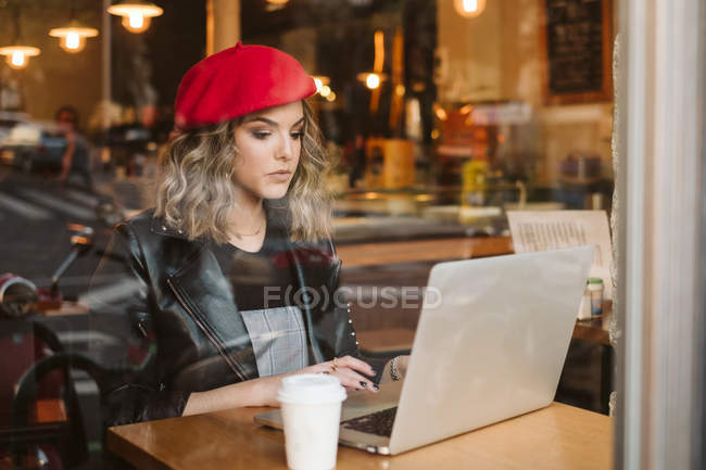 Молода жінка в червоному береті з гарячим напоєм і ноутбуком, сидячи в кафе — стокове фото