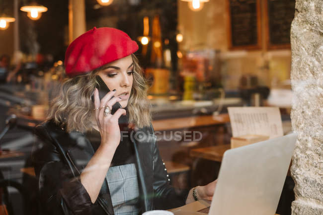Mujer joven de moda en boina roja en el teléfono mientras está sentado en la mesa con el ordenador portátil en el restaurante - foto de stock