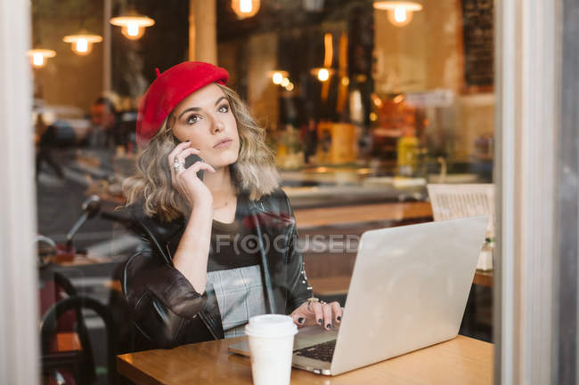 Модная молодая женщина в красном берет по телефону, сидя за столом с ноутбуком в ресторане — стоковое фото