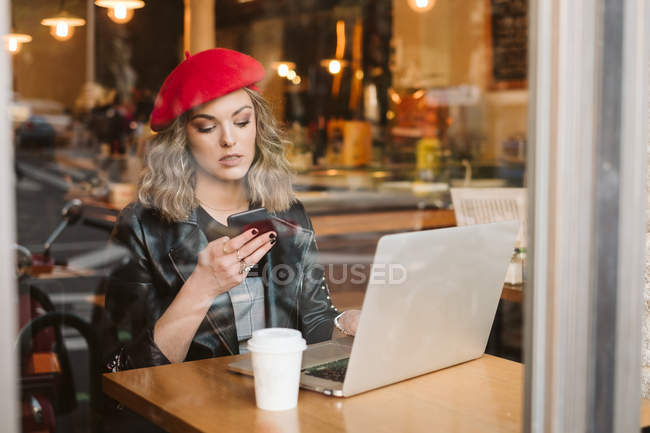 Trendige junge Frau in roter Baskenmütze mit Handy, während sie mit Laptop am Tisch im Restaurant sitzt — Stockfoto