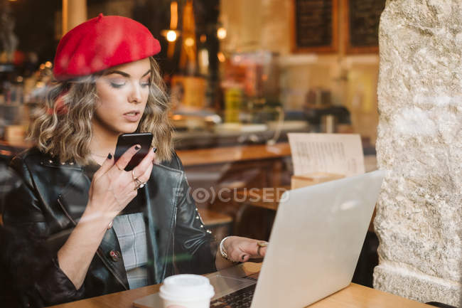 Menina na moda em boina vermelha usando telefone celular enquanto sentado à mesa com laptop no restaurante — Fotografia de Stock
