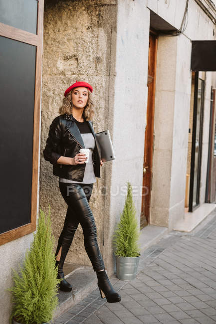 Elegante joven hembra en boina roja sosteniendo bebida para llevar mientras sale de la cafetería en la calle de la ciudad - foto de stock