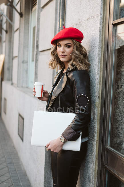 Stylish young female in red beret holding takeaway beverage leaning against wall of a coffee shop on city street — Stock Photo