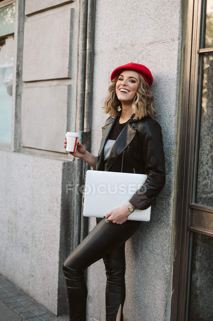 Stylish laughing young female in red beret holding takeaway beverage leaning against wall of a coffee shop on city street — Stock Photo