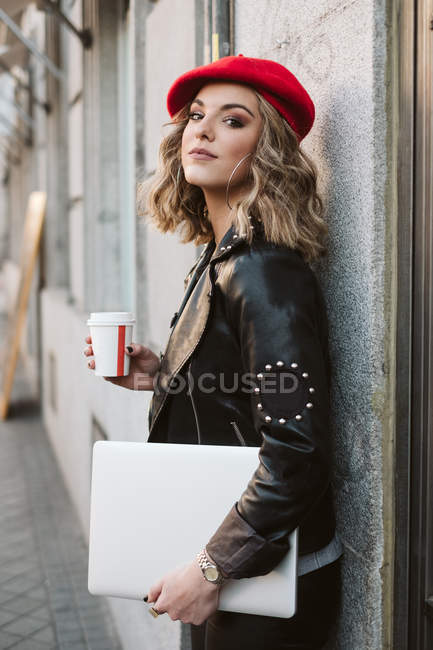 Stylish young female in red beret holding takeaway beverage leaning against wall of a coffee shop on city street — Stock Photo