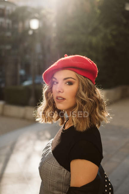 Jeune femme portant béret rouge à la mode regardant la caméra tout en se tenant sur la rue de la ville par jour ensoleillé — Photo de stock