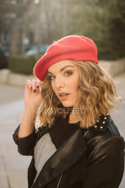 Молода жінка в модному червоному береті дивиться на камеру, стоячи на вулиці міста в сонячний день — стокове фото