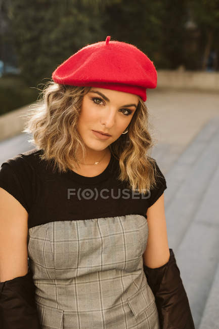Молода жінка в модному червоному береті дивиться на камеру, стоячи на вулиці міста в сонячний день — стокове фото