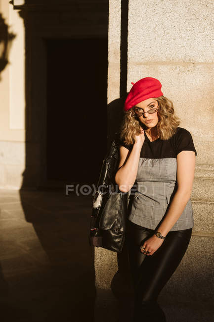 Sensuale giovane donna in berretto alla moda e occhiali tenendo giacca di pelle sulla spalla e guardando la fotocamera mentre in piedi vicino al muro edificio nella giornata di sole sulla strada della città — Foto stock