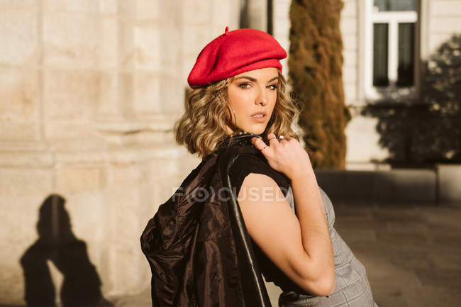 Sinnliche junge Frau in trendiger Baskenmütze mit Lederjacke auf der Schulter und Blick in die Kamera, während sie an einem sonnigen Tag an der Stadtstraße in der Nähe einer Hauswand steht — Stockfoto