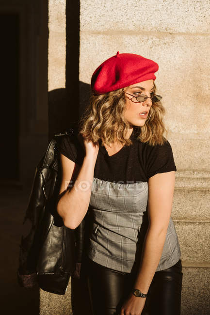 Чуттєва молода жінка в модному береті і окулярах тримає шкіряну куртку на плечі і дивиться геть, стоячи біля будівельної стіни в сонячний день на вулиці міста — стокове фото