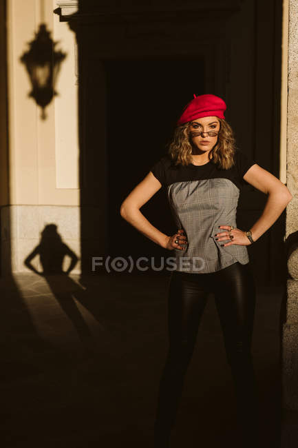 Уверенная в себе молодая женщина в модном берете и очках смотрит в камеру, стоя возле стены здания в солнечный день на городской улице — стоковое фото