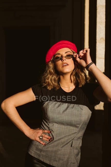 Чувственная молодая женщина в модном берете и очках смотрит в камеру, стоя возле стены здания в солнечный день на городской улице — стоковое фото