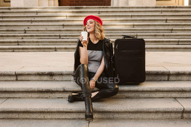 Щаслива молода жінка в модному вбранні, дивлячись далеко, сидячи на сходах біля валізи і насолоджуючись виносним напоєм — стокове фото