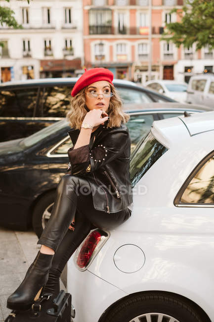 Junge Frau im trendigen Outfit tritt auf Koffer, während sie sich an Auto lehnt — Stockfoto