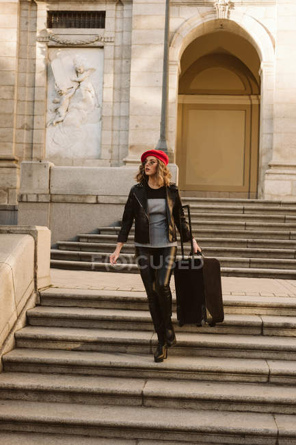 Молодая женщина в модном наряде в современных солнцезащитных очках идет по лестнице с чемоданом — стоковое фото