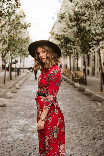 Чуттєва молода жінка в стильному одязі і капелюсі дивиться на камеру, стоячи на старій тротуарі на міській вулиці — стокове фото