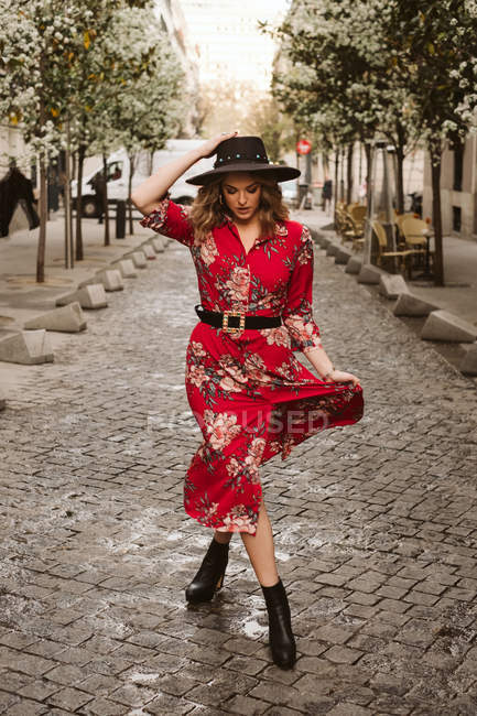 Young woman in stylish dress and hat walking on aged pavement on city street — Stock Photo