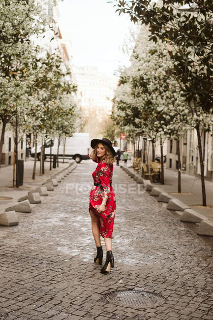 Rückansicht einer jungen Frau in stylischem Kleid und Hut, die der Kamera über die Schulter schaut, während sie auf dem Bürgersteig der Stadtstraße spaziert — Stockfoto