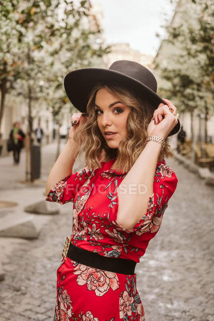 Sinnliche junge Frau in stylischem Kleid und Hut, die in die Kamera blickt, während sie auf dem Bürgersteig der Stadtstraße steht — Stockfoto