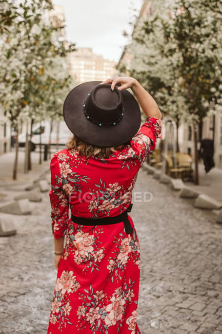 Vue arrière de la jeune femme en robe élégante et chapeau marchant sur le trottoir vieilli sur la rue de la ville — Photo de stock