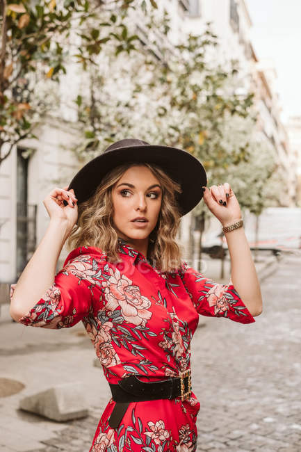 Jeune femme sensuelle en robe élégante et chapeau regardant la caméra tout en se tenant debout sur le trottoir âgé sur la rue de la ville — Photo de stock