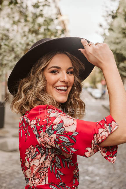 Rindo jovem mulher em vestido elegante e chapéu olhando para a câmera no pavimento envelhecido na rua da cidade — Fotografia de Stock