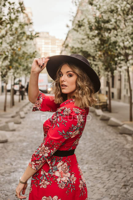 Sensual jovem mulher em vestido elegante e chapéu olhando para a câmera enquanto estava em pé no pavimento envelhecido na rua da cidade — Fotografia de Stock