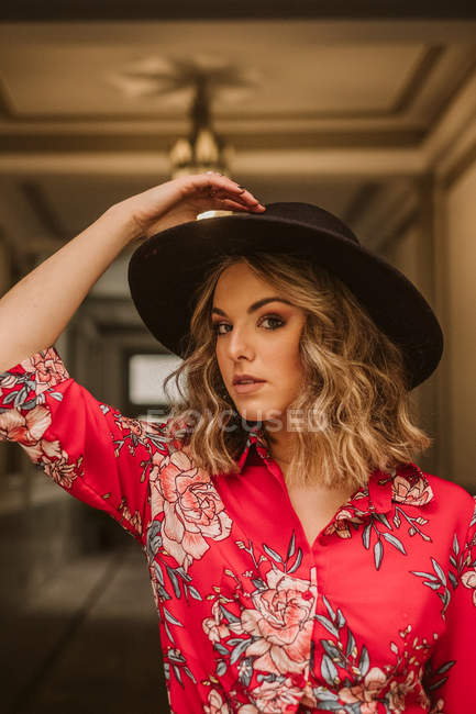 Стильна молода жінка в елегантній сукні і капелюсі дивиться на камеру, стоячи під лампою в проходженні старої будівлі — стокове фото