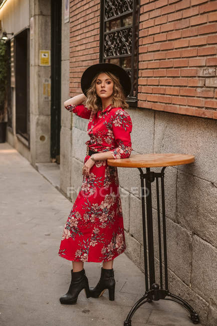 Trendy young female in elegant dress and hat looking away while leaning on round table near old building on city street — Stock Photo