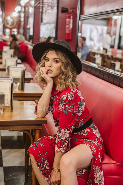 Чуттєва молода жінка в стильній елегантній сукні і капелюсі сидить на червоному дивані біля столу і дивиться на камеру в ресторані — стокове фото