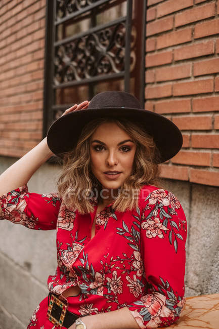 Attraktive junge Frau in stylischem Kleid und Hut blickt in die Kamera in der Nähe eines alten Gebäudes an der Stadtstraße — Stockfoto