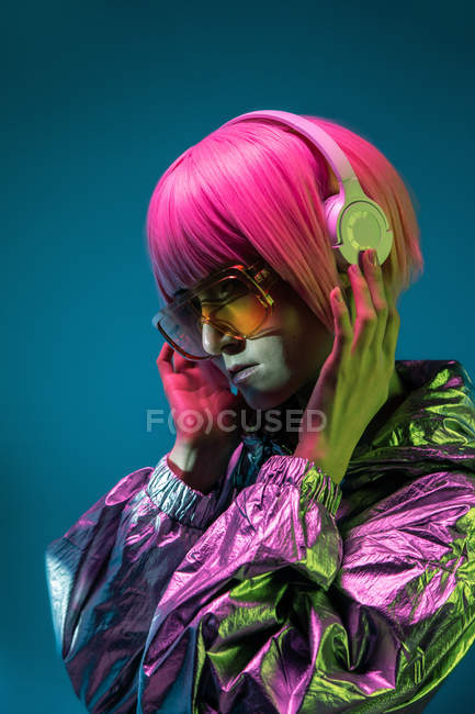 Mujer asiática joven con elegante corte de pelo rosa y chaqueta de plata brillante de pie y escuchar música - foto de stock
