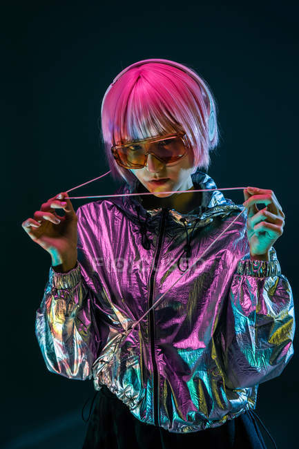 Trendy young Japanese woman with purple hair standing in sparkly silver jacket and red sunglasses on blue background — Stock Photo