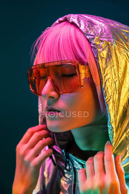 Trendy young Japanese woman with purple hair standing in silver hoodie jacket and red sunglasses on blue background — Stock Photo