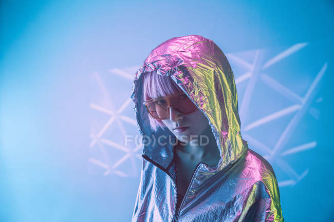 Trendy young Japanese woman with purple hair standing in silver hoodie jacket and red sunglasses on blue background — Stock Photo