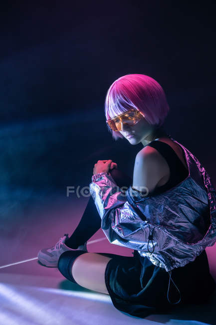 Trendy young Japanese woman with purple hair in silver jacket and red sunglasses sitting on dark background — Stock Photo
