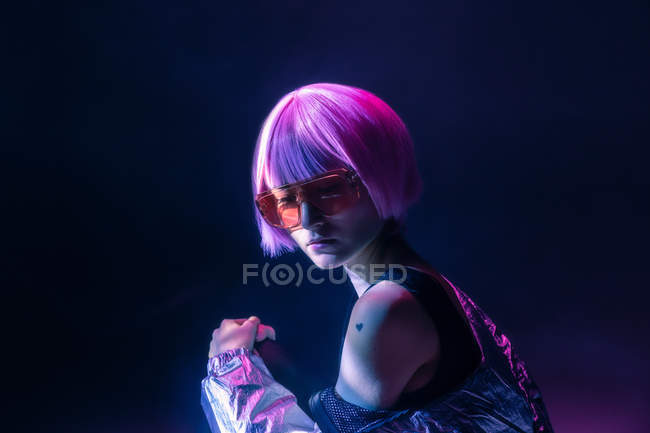 Trendy young Japanese woman with purple hair in sparkly silver jacket and red sunglasses posing on dark background — Stock Photo